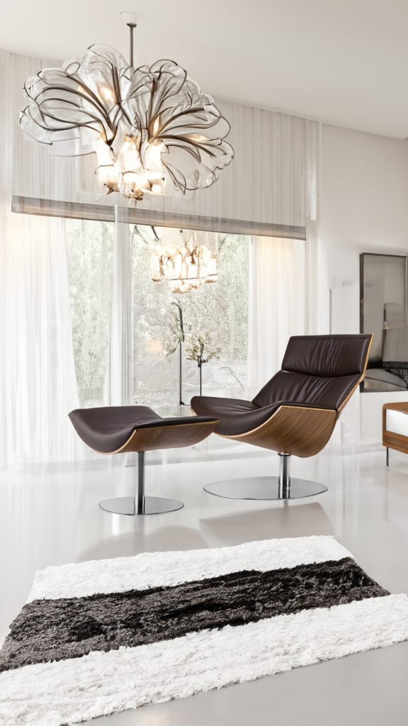Szary salon nowoczesny - inspiracja. Na tle drzwi balkonowych widnieje duży fotel wypoczynkowy  z podnóżkiem ustawiony obok białego dywanu z brązowym paskiem. Pomieszczenie doświetla ozdobny żyrandol. 